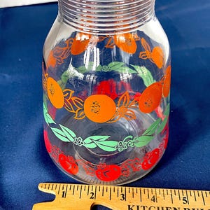 May include: A clear glass pitcher with a wide base and a narrow neck. The pitcher is decorated with orange fruit and green leaf designs. A wooden ruler with the text "KITCHEN RULE" is in the foreground.