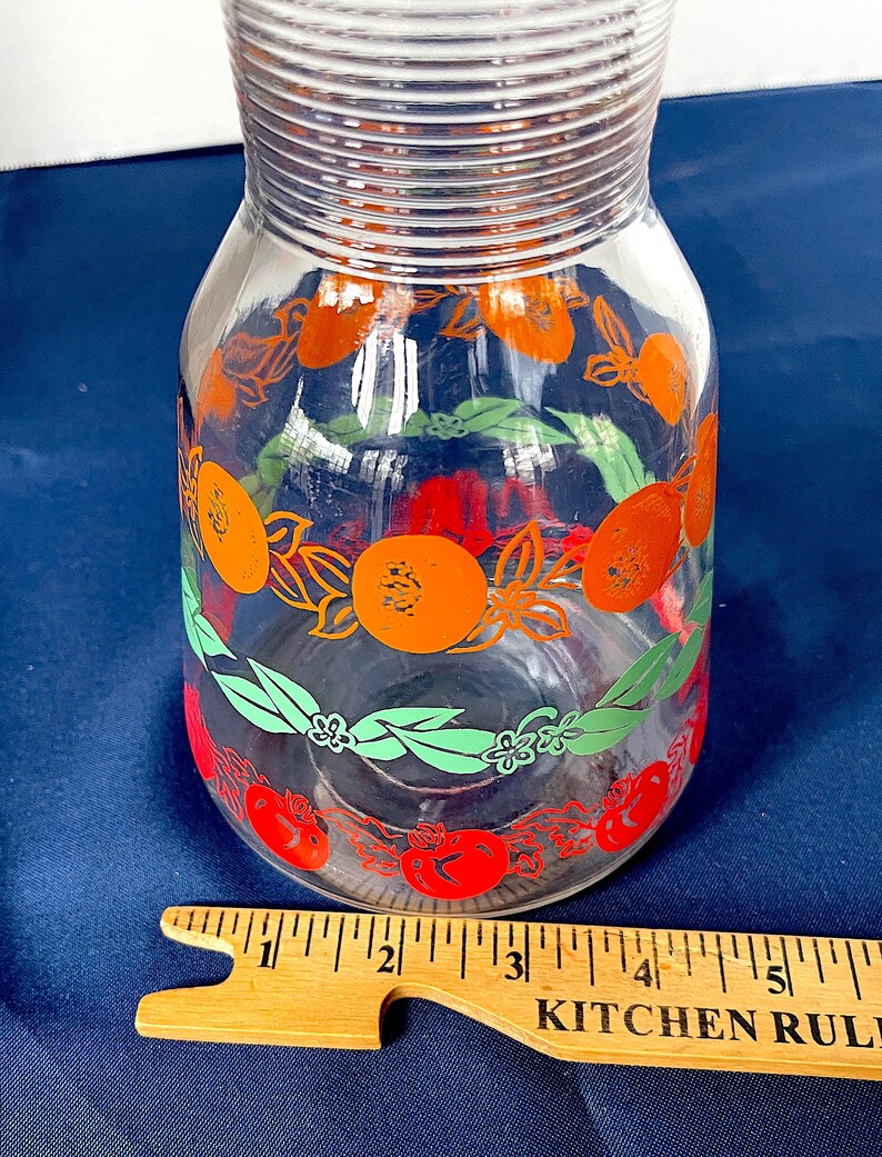 May include: A clear glass pitcher with a wide base and a narrow neck. The pitcher is decorated with orange fruit and green leaf designs. A wooden ruler with the text "KITCHEN RULE" is in the foreground.