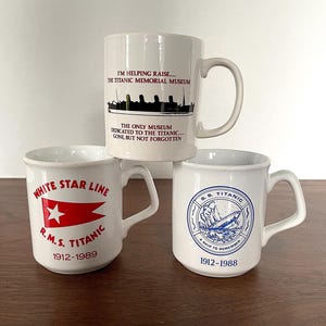 May include: Three white ceramic mugs with Titanic-themed designs. One mug features the text "I'M HELPING RAISE... THE TITANIC MEMORIAL MUSEUM". Another displays the White Star Line logo and "R.M.S. TITANIC 1912-1989". The third mug has a blue emblem and "S.S. TITANIC 1912-1988".