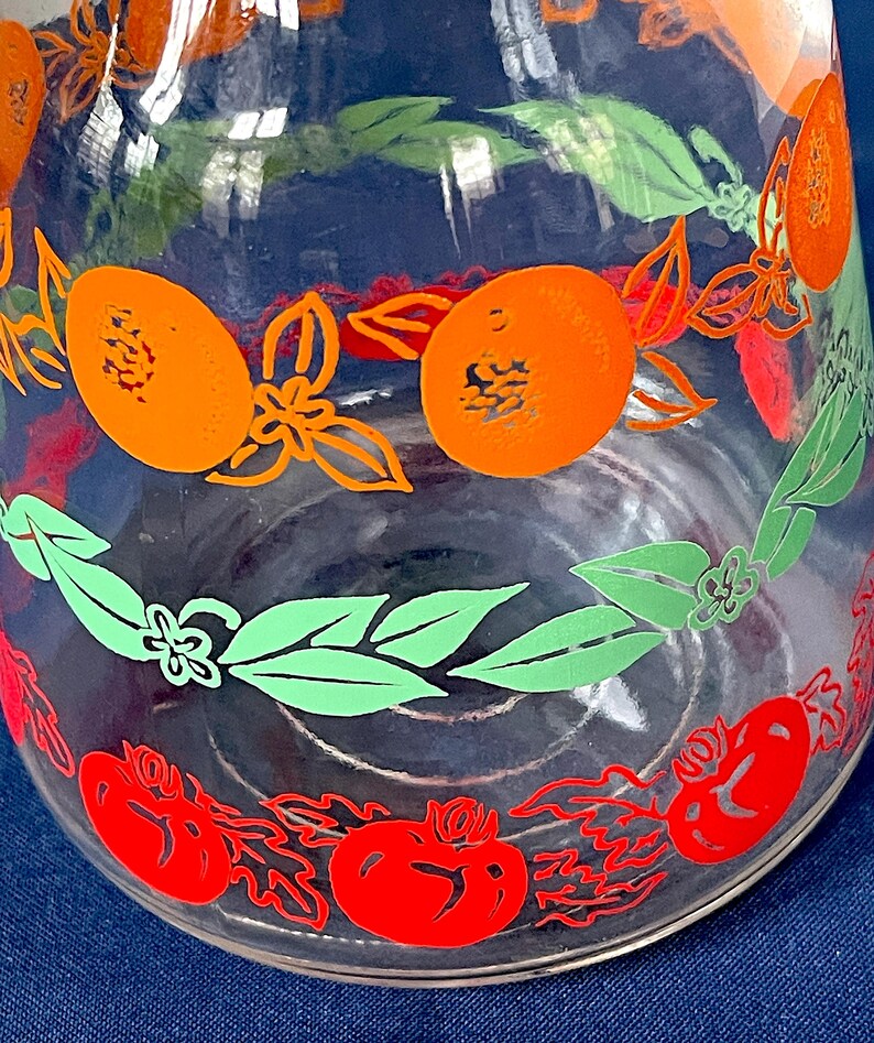 May include: Clear glass pitcher with a colorful fruit design. The pitcher features orange, red, and green fruit and leaf patterns. The design includes oranges, tomatoes, and leaves, creating a vibrant and decorative look. Ideal for serving beverages.