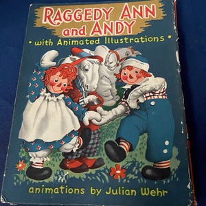 May include: Vintage children's book titled "Raggedy Ann and Andy" with animated illustrations. The cover features illustrations of Raggedy Ann, Andy, and a white horse. The book has a blue background with text in red and white.