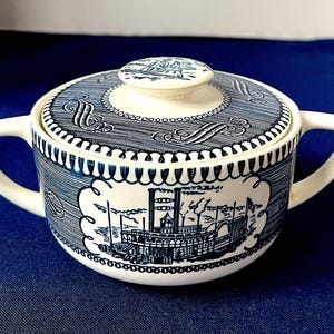 Może przedstawiać: Biało-niebieska ceramiczna cukiernica z pokrywką. Miska przedstawia szczegółową ilustrację parowca i dekoracyjne wzory. Pokrywka ma mały uchwyt i pasujący wzór. Miska ma dwa uchwyty.