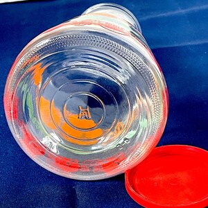 May include: A clear glass jar with a red lid, lying on its side. The jar has colorful floral designs in red, orange, and green. The lid is a solid, bright red. The bottom of the jar has a stamped logo.
