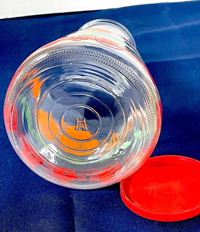 May include: A clear glass jar with a red lid, lying on its side. The jar has colorful floral designs in red, orange, and green. The lid is a solid, bright red. The bottom of the jar has a stamped logo.