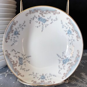 May include: A white porcelain soup bowl with a gold rim and a delicate floral pattern in shades of blue and brown. The bowl features a repeating design of stylized roses and leaves.