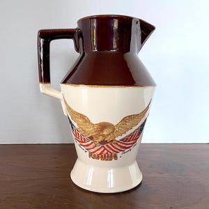 Może przedstawiać: Dzbanek ceramiczny w kolorze brązowym i białym z wzorem orła trzymającego amerykańskie flagi. Na dzbanku widnieje napis "The Spirit of" i "American Pride".