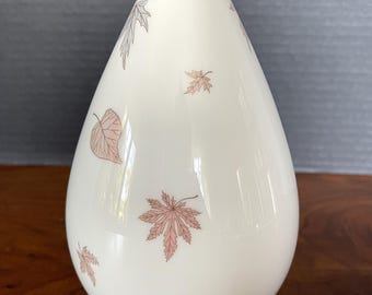 May include: A white ceramic vase with a teardrop shape and a gold rim. The vase is decorated with a pattern of pink and gray leaves.