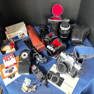 May include: A selection of vintage photography equipment, including a Minolta SRT-101 camera, lenses, flashbulbs, and manuals. The items are arranged on a blue surface, with various cases and accessories visible.