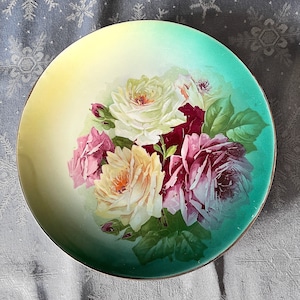 Puede incluir: Un plato de porcelana vintage con un diseño pintado a mano de un ramo de rosas en tonos de rosa, blanco y rojo. Las rosas están rodeadas de hojas verdes y se colocan sobre un fondo verde y amarillo.