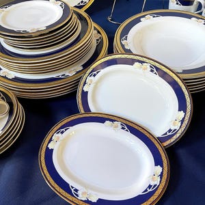 May include: A collection of fine china with a white center, navy blue border, and gold trim. The set includes plates, bowls, and serving platters. Floral accents decorate the navy border. The china is arranged on a dark blue surface.