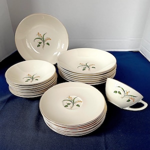 May include: A collection of cream-colored vintage dishware with a gold rim and a floral design. The set includes dinner plates, saucers, bowls, and a teacup. The floral design features green leaves and yellow flowers.