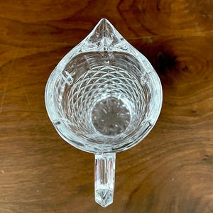 Op de afbeelding: Een glazen kan met een diamantpatroon en een handvat. De kan is leeg en staat op een houten oppervlak.