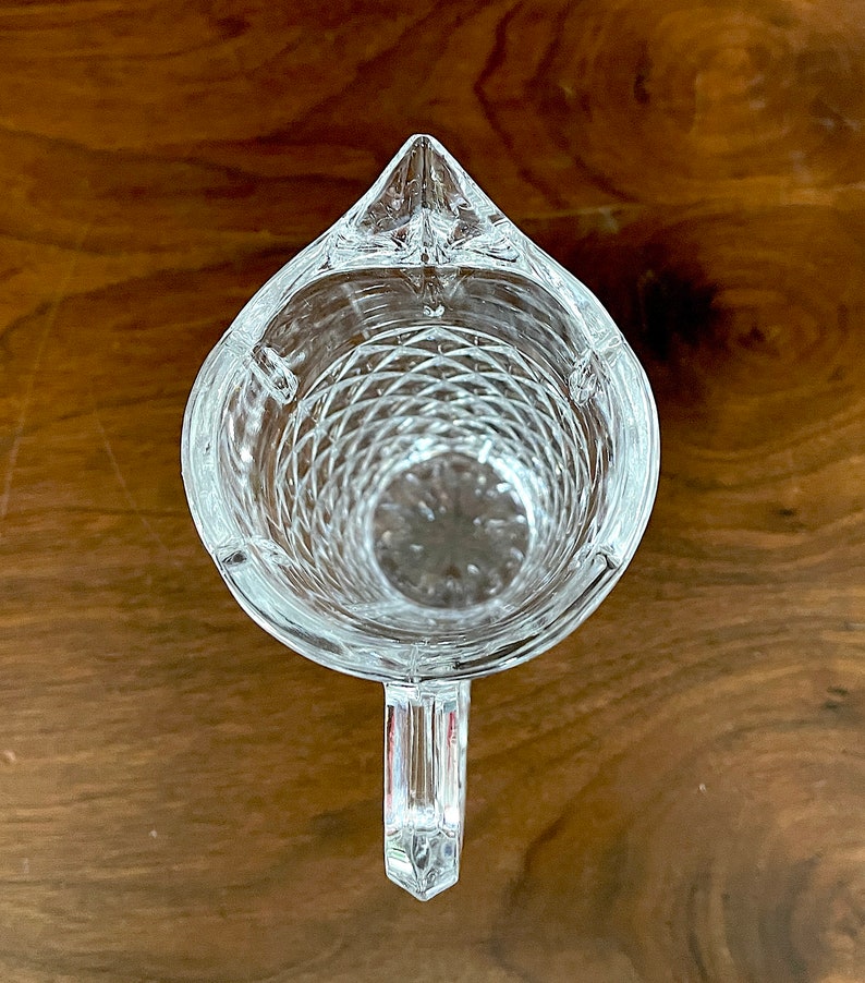 Op de afbeelding: Een glazen kan met een diamantpatroon en een handvat. De kan is leeg en staat op een houten oppervlak.