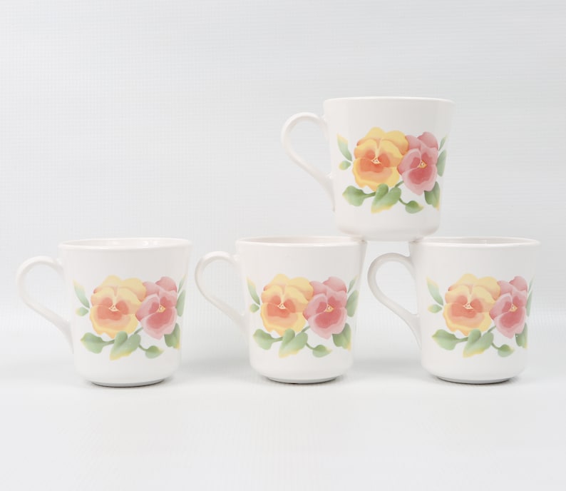 May include: Set of four white ceramic mugs with a floral design. Each mug features a yellow and pink pansy flower with green leaves.