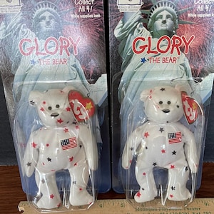 Könnte beinhalten: Zwei weiße Beanie Baby Bären mit roten, weißen und blauen Sternen und Streifen. Die Bären sind in Plastikbehältern mit einem Hintergrundbild der Freiheitsstatue verpackt. Die Verpackung lautet "Collect All 4!" und "Glory the Bear".