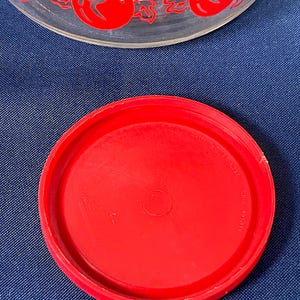 May include: A clear glass bowl with a red tomato and leaf design around the rim. A bright red plastic lid is in the foreground. The lid is circular and has a slightly raised edge.