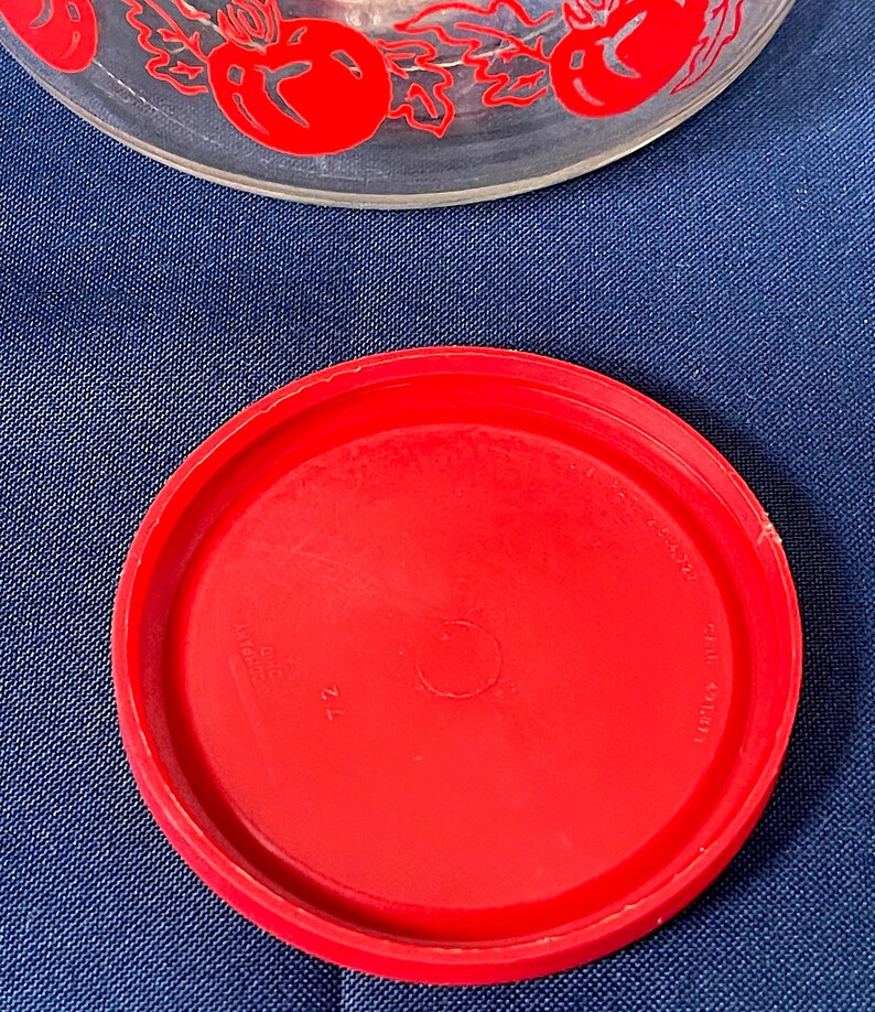 May include: A clear glass bowl with a red tomato and leaf design around the rim. A bright red plastic lid is in the foreground. The lid is circular and has a slightly raised edge.
