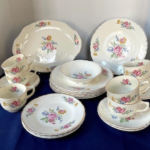 Puede incluir: Un juego de porcelana crema vintage con diseños florales con rosas rosas, amarillas y azules. El juego incluye platos, cuencos, platillos y tazas de té con ribetes dorados, dispuestos sobre una superficie azul.