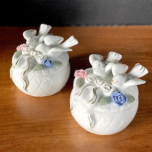 May include: Two white porcelain trinket boxes with lids, each featuring a pair of sculpted white birds, a gold-trimmed bow, and small pink and blue floral accents. The base of each round container has a raised diamond-patterned texture.