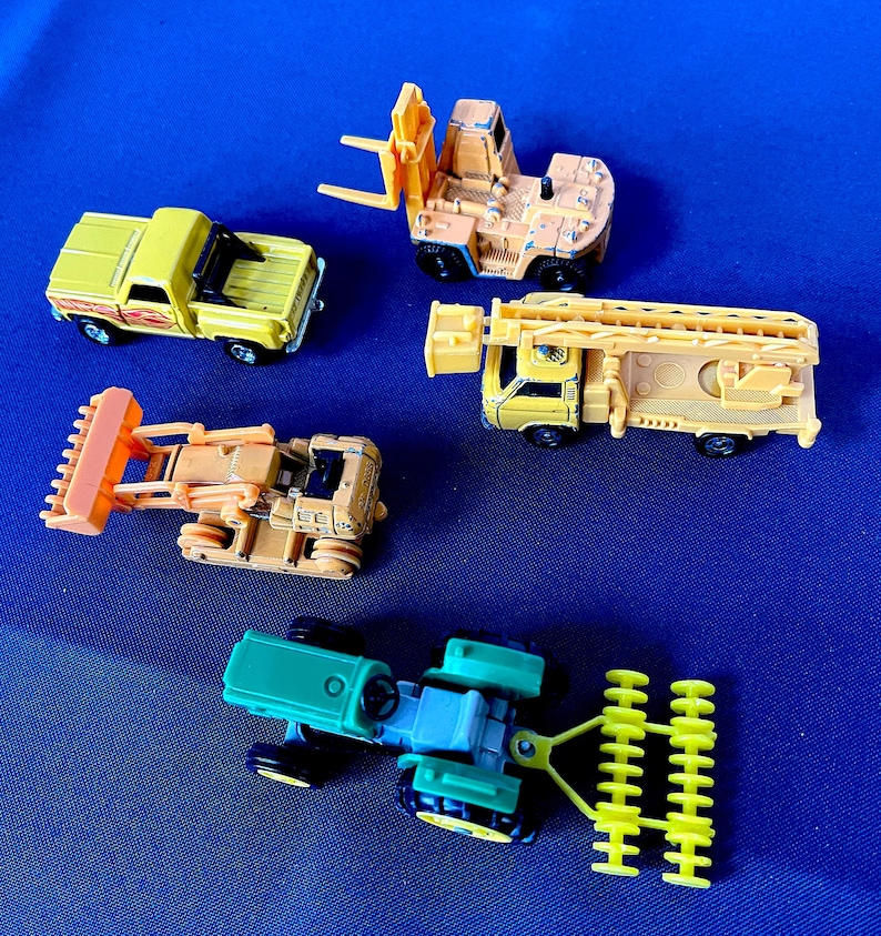 May include: A collection of toy construction vehicles, including a yellow pickup truck, a forklift, a bulldozer, a fire truck, and a tractor. The toys are in various shades of yellow, orange, and green, set against a blue background.