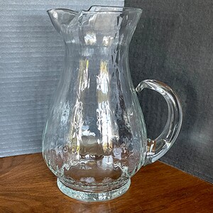 May include: A clear glass pitcher with a textured surface and a handle. The pitcher has a wide base and a narrow neck.