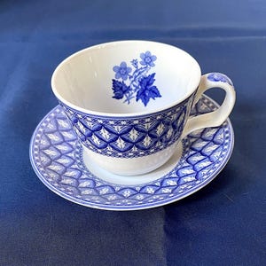 May include: White teacup and saucer set with a blue floral design inside the cup and a geometric pattern on the cup's exterior and saucer. The handle of the cup also has a blue floral design. The set is on a blue surface.