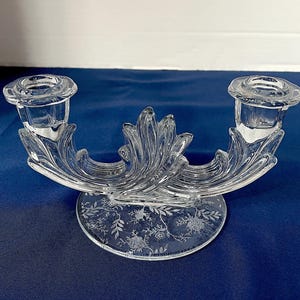 May include: Clear glass candle holder with two candle cups. The candle holder has a decorative, curved design with leaf-like details. The base is round with an etched floral pattern. The candle holder is on a blue surface.