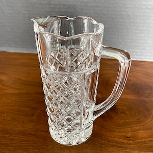 Op de afbeelding: Een heldere glazen kan met een diamantpatroon en een gegolfde rand. De kan heeft een handvat en staat op een houten oppervlak.