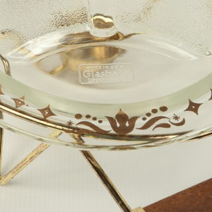 May include: A clear glass serving dish with a gold rim and gold floral design. The dish is on a gold metal stand with three legs. The dish has the text "Made in USA Glasbake 1920" on the bottom.
