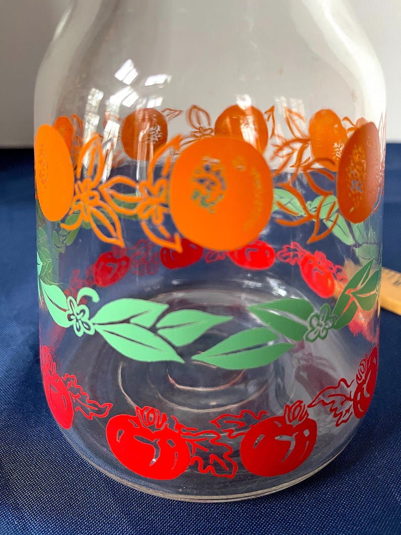 May include: Clear glass pitcher with a decorative fruit design. The pitcher features orange, red, and green illustrations of oranges, tomatoes, and leaves. The design wraps around the pitcher, creating a vibrant and colorful aesthetic.