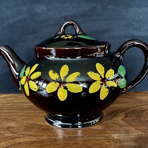 Op de afbeelding: Een bruine theepot met een bloemenpatroon. De theepot heeft een rond lichaam met een korte tuit en een handvat. Het bloemenpatroon heeft gele bloemen met rode centra en groene bladeren op een zwarte achtergrond.
