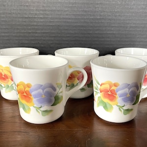 May include: Five white ceramic mugs with a floral design featuring yellow, orange, and purple pansies. The mugs have a rounded shape and a handle on the side.