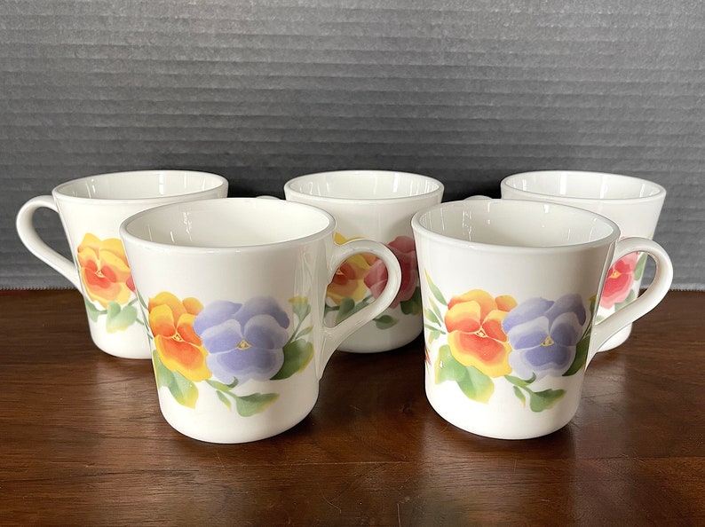 May include: Five white ceramic mugs with a floral design featuring yellow, orange, and purple pansies. The mugs have a rounded shape and a handle on the side.