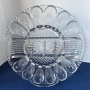 May include: A clear, glass serving dish with a divided design. The dish features a circular shape with a scalloped edge and multiple compartments. The glass has a textured, cut-glass pattern, adding visual interest to the piece.