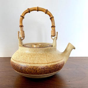 May include: A brown and beige ceramic teapot with a bamboo handle. The teapot has a speckled glaze and a brown stripe around the middle.