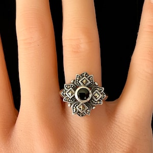 Puede incluir: Un anillo de plata con una piedra preciosa negra rodeada de un diseño detallado de piedras de marcasita.