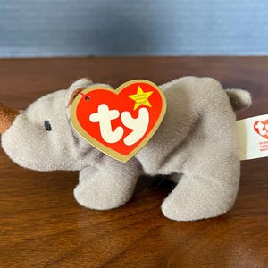 May include: A gray plush rhinoceros with a brown horn and a red Ty heart tag with the word "ty" in white. The tag also has a yellow star. The rhinoceros is sitting on a wooden surface.