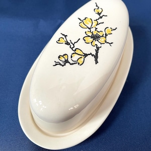 Könnte beinhalten: Weißer Keramik-Butterbehälter mit Deckel. Der Deckel ist mit einem gelben Blumendesign mit schwarzen Stielen und Akzenten verziert. Das Gericht hat eine ovale Form und steht auf einem passenden ovalen Sockel. Der Hintergrund ist einfarbig blau.