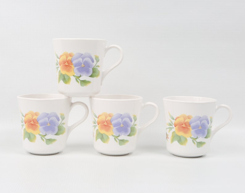 May include: Four white ceramic mugs with a floral design featuring yellow and purple pansies. The mugs have a white background and a simple handle.