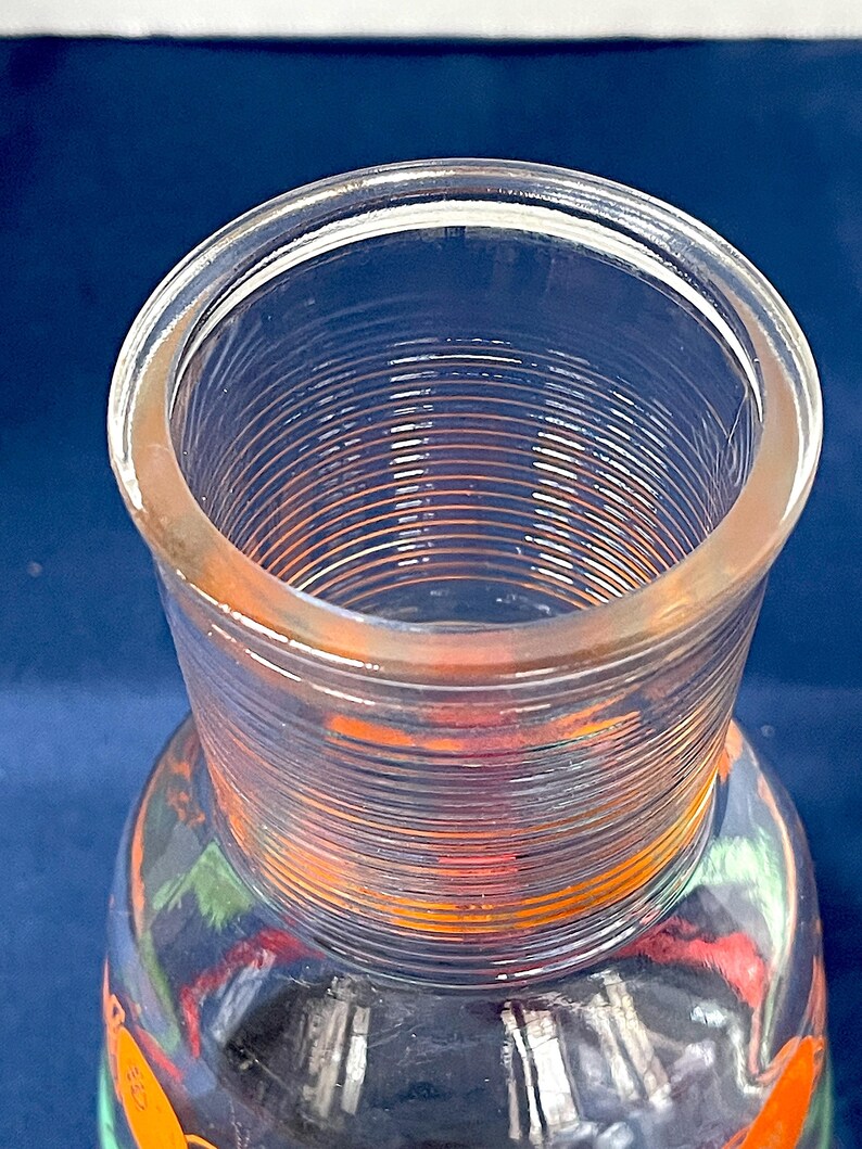 May include: Clear glass pitcher with a wide, ribbed neck and a colorful design on the lower portion. The pitcher has a slightly flared rim and a cylindrical neck. The design on the pitcher includes orange and green elements.