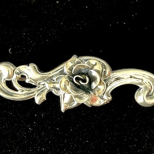 May include: A silver brooch in the shape of a stylized flower with a swirl design. The flower has a raised center and is surrounded by delicate, swirling leaves.