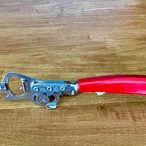 May include: A manual can opener featuring a bright red plastic handle and a silver-colored metal mechanism. The metal body includes a cutting wheel, a gear, and a bottle opener notch, with small embossed text visible on the side of the metal frame.