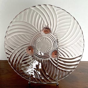 May include: A clear glass serving bowl with a swirl pattern and three pink glass feet. The bowl is on a wooden surface.
