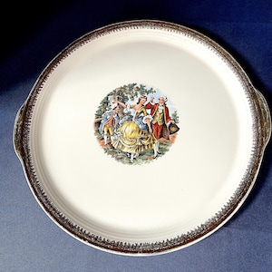 Könnte beinhalten: Ein runder, cremefarbener Servierteller mit goldfarbenem Rand und Griffen. In der Mitte befindet sich eine farbenfrohe Illustration von Menschen in Kleidung aus dem 18. Jahrhundert. Der Rand hat ein detailliertes, strukturiertes Design. Ein dekoratives Servierstück.