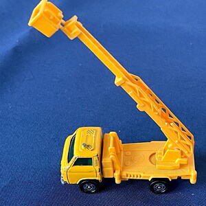 May include: A yellow toy truck with a crane arm extended upwards. The truck has black wheels and a detailed design, including a ladder and a small bucket at the end of the crane. The toy is set against a blue background.