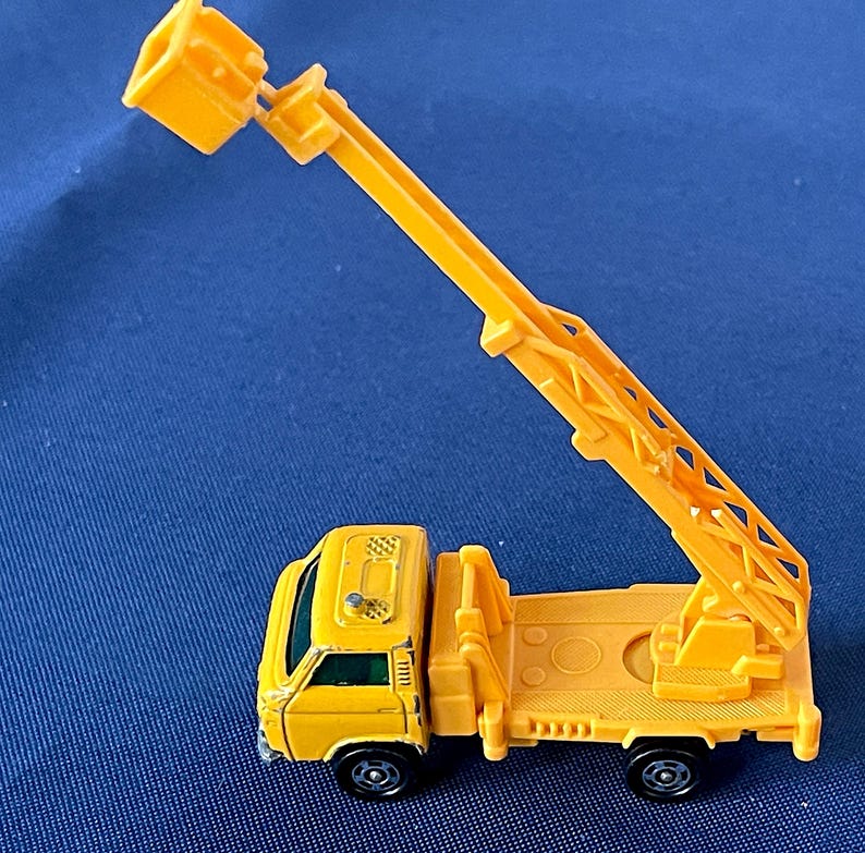 May include: A yellow toy truck with a crane arm extended upwards. The truck has black wheels and a detailed design, including a ladder and a small bucket at the end of the crane. The toy is set against a blue background.