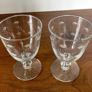 May include: Two clear glass goblets with etched leaf and stem designs. The goblets have a rounded bowl shape, a short stem, and a circular base. The etched design features small leaves and delicate stems around the bowl.