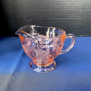 May include: A pink glass creamer with a floral pattern. The creamer has a handle and a spout for pouring. The base is round and the body is decorated with embossed flowers. The creamer is sitting on a blue surface.