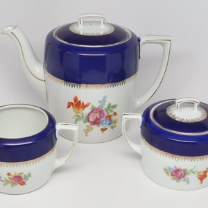 Puede incluir: Un juego de té de porcelana blanca con detalles azules y dorados. La tetera, la jarra de crema y el azucarero presentan un diseño floral con una banda azul alrededor de la parte superior.