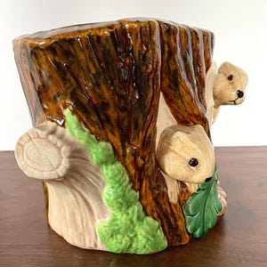 May include: A ceramic tree stump planter with two squirrels. The planter is painted in brown, tan, and green, with a glossy finish. The squirrels have brown fur and black eyes. The planter is designed to hold plants.
